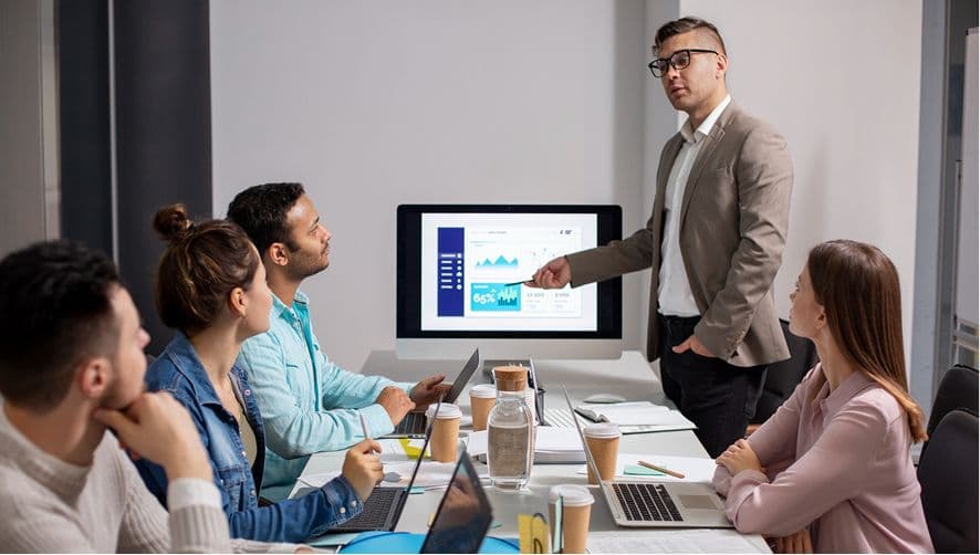 Man presents business data with graphs on a screen to five colleagues in a meeting.